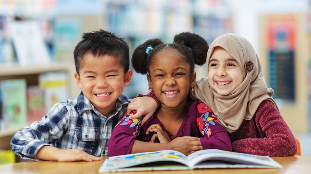 Three young children smiling with an open picture book on the table in front of them. The child on the left is a young boy with tan skin and black hair wearing a blue plaid shirt. The child in the middle is a young girl with brown skin and dark brown hair in buns wearing a purple shirt with flowers. The child on the right is a young girl with light skin wearing a tan hijab and a dark pink sweater.