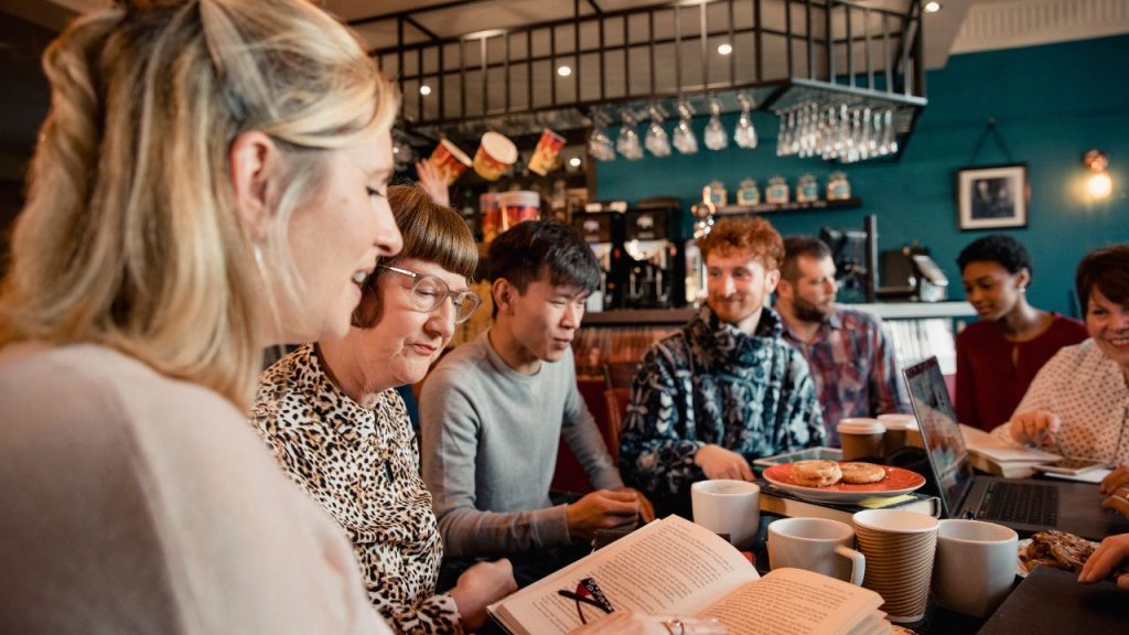 Seven people of varying ages, genders, and ethnicities sitting at at a table in a busy coffeeshop. Many are talking to one another and smiling, and there are open books, coffee mugs, and pastries crowded on the table.