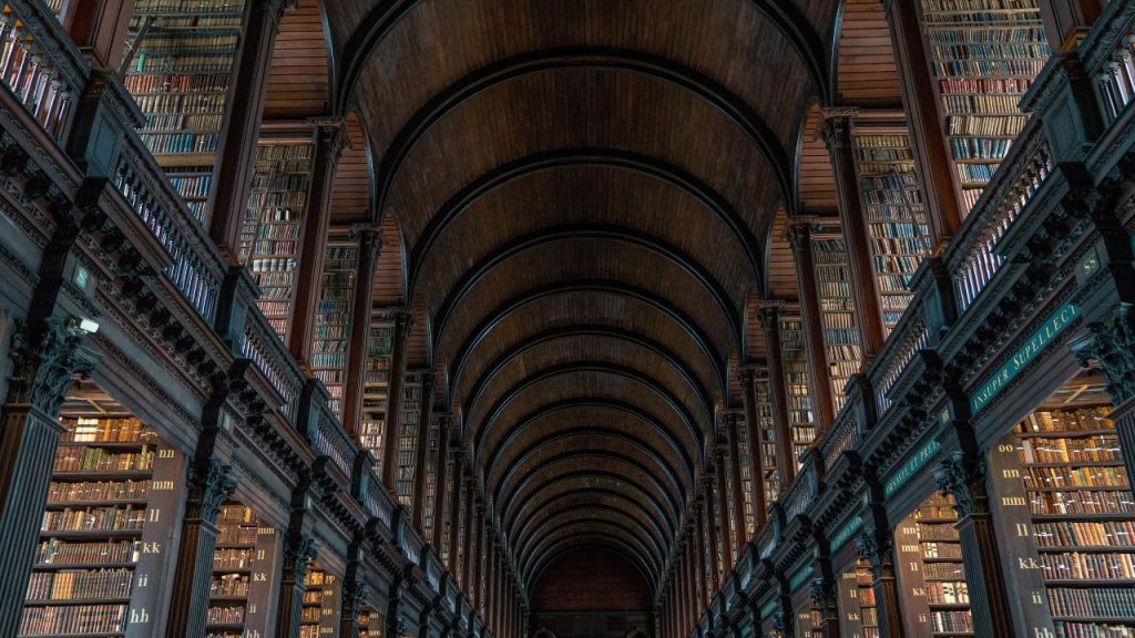 A vast historic library hallway with a curved wooden ceiling and dark brown balconies. Rows upon rows of book spines sit on tall shelves that span at least the height of two floors.