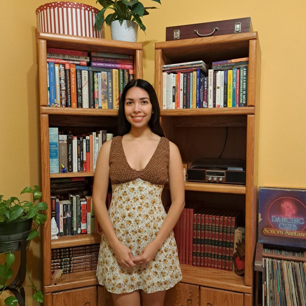 Photo of Anamely standing in front of her bookshelves.