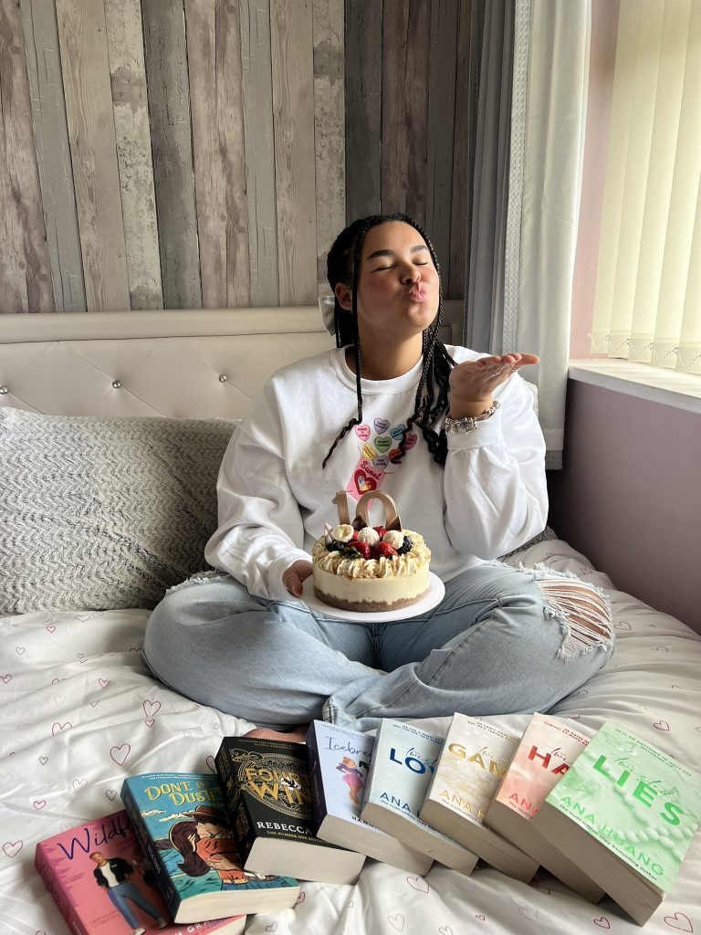 Tia sitting on a soft bed, holding a yummy dessert. There is a fan of books laid out on the bed before her.