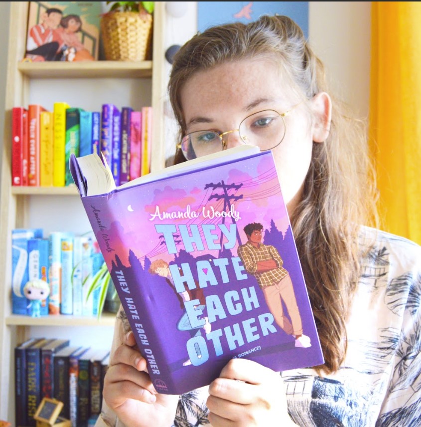 Eloise is shown sitting before her bookshelf in a well lit room, reading Amanda Woody's They Hate Each Other.