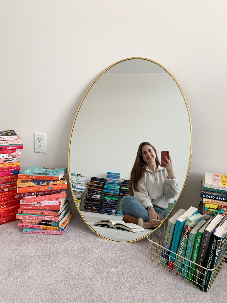 Photo of Melissa's room. She is featured in the mirror leaning on the wall. There are piles of books everywhere.