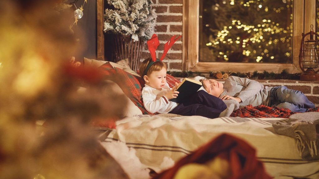 Two young kids relax together on a bed with holiday decorations surrounding them. One is wearing red reindeer antlers and reading a book, while the other sleeps near him. There are holiday lights on bushes outside a large window in the background.