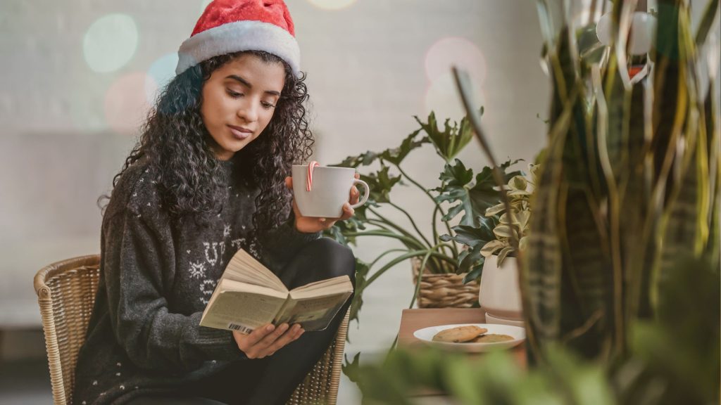 A woman reads a book at a table while drinking a warm beverage with a candy cane in a white mug. She has long, curly black hair and is wearing a Santa hat. On the table in front of her is a plate of cookies and green plants decorate the room.