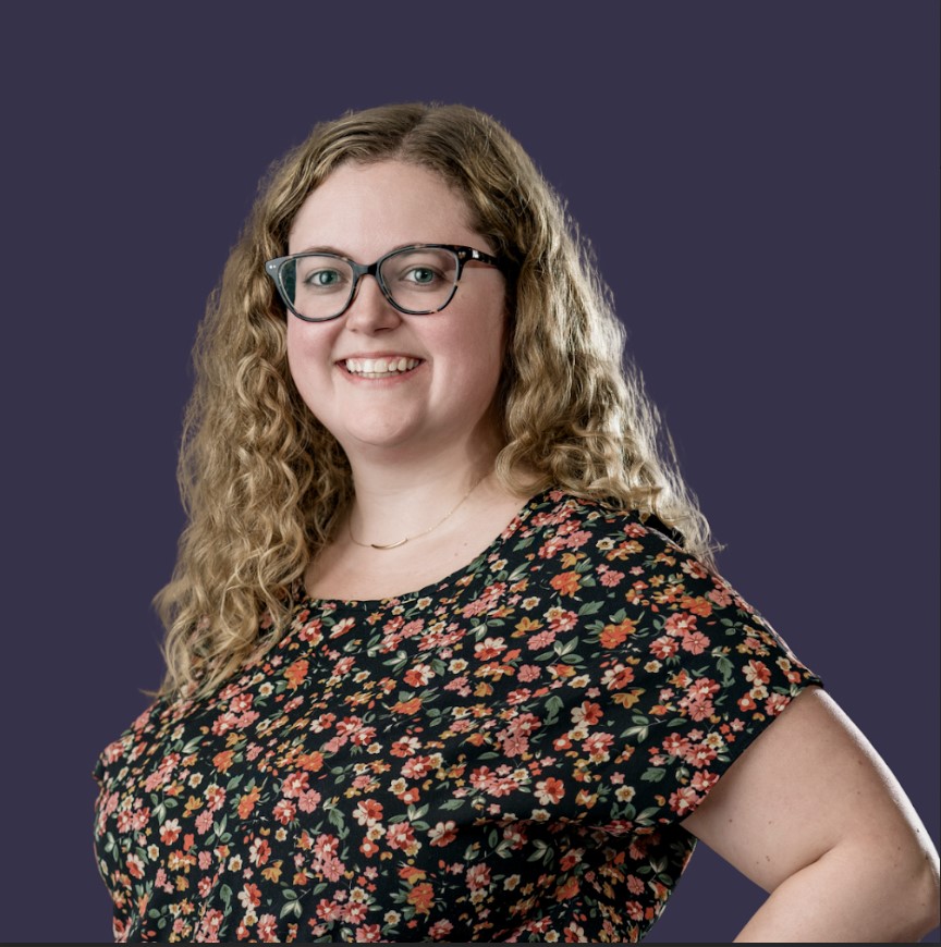 Kayla Martin smiling, wearing a floral top and standing against a blue background.