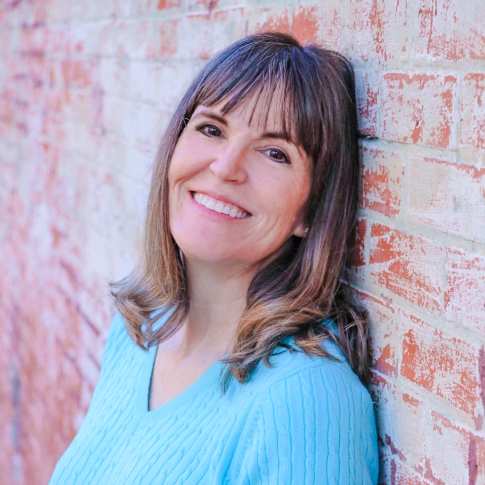 Tamie Dearen is smiling, wearing a blue sweater, and leaning against a brick wall.