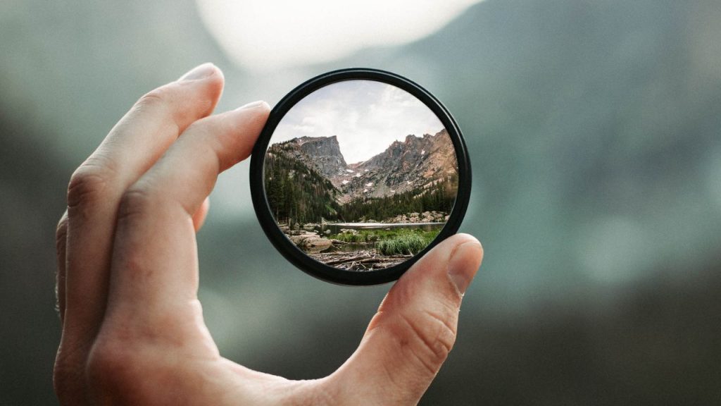 Over a blurry background, a hand holds a black framed circle that shows a high resolution mountain landscape
