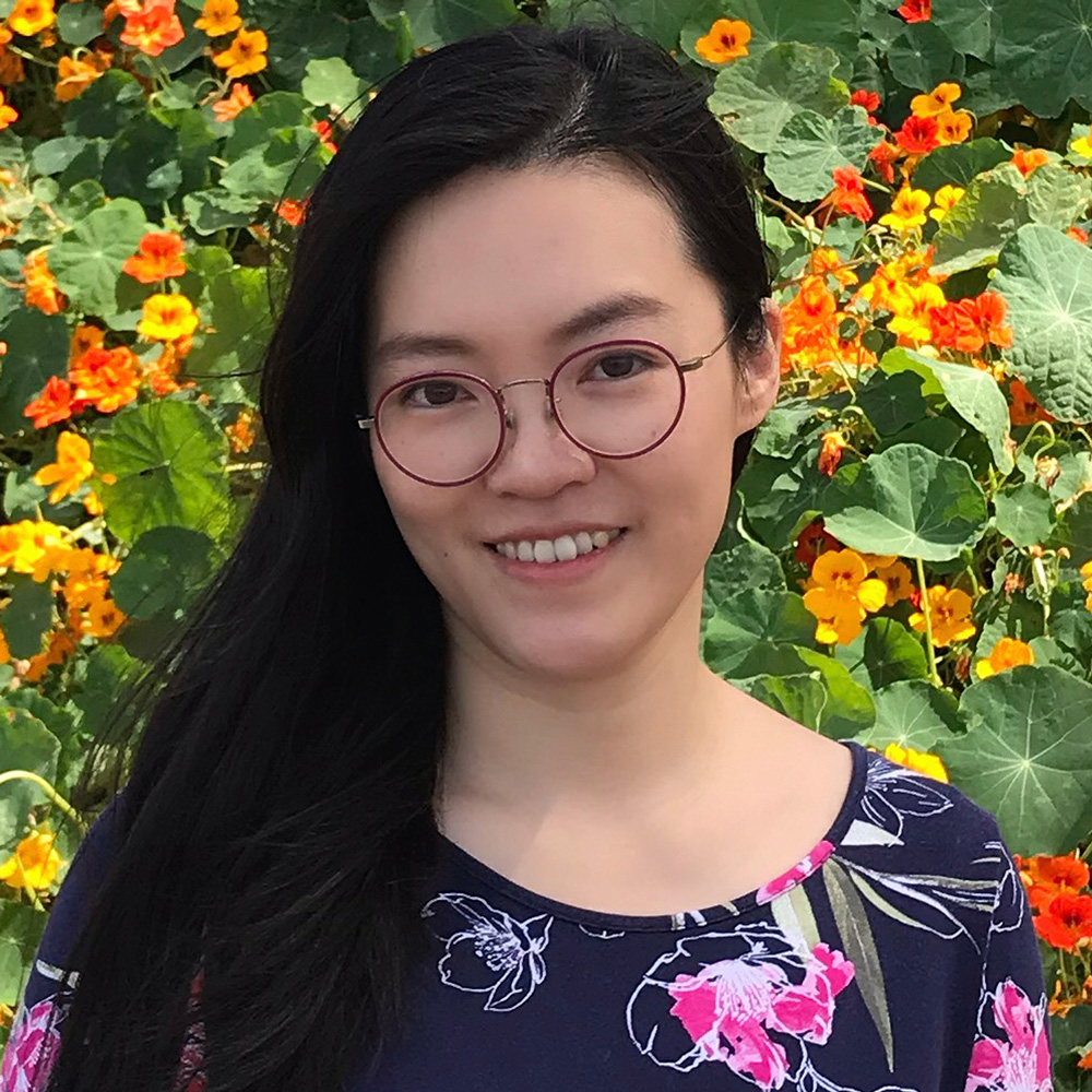Helen H. Wu, a pale-skinned, black-haired east-Asian woman wearing red-rimmed eyeglasses and a navy blue-and-pink flowered blouse smiles at the viewer; in the background is are brightly-colored orange flowers among large green leaves.