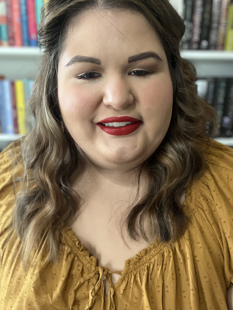 Photo of author Tati B. Alvarez. She wears a yellow shirt and is smiling at the camera with her bookshelves behind her.