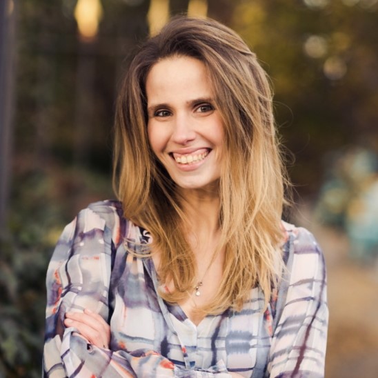 A white woman with long balayage brown/blonde hair and wearing a blue-and-white plaid blouse smiles at the viewer; in the background is a forested area.