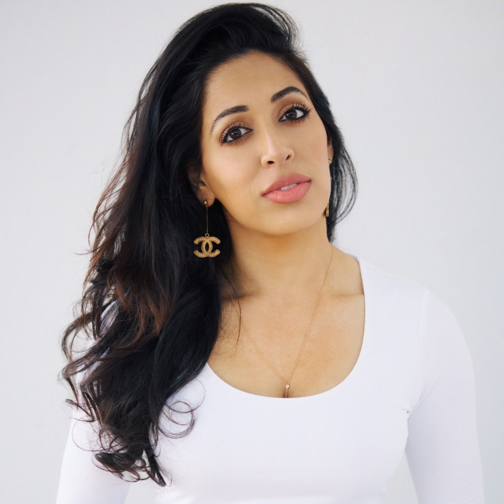 A brown-skinned woman with long black hair and wearing a white blouse and Chanel earrings smiles at the viewer; the background is a white backdrop.