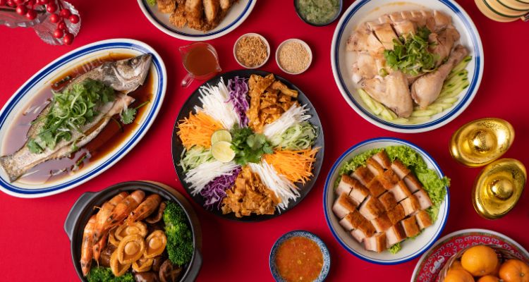An overhead view of a traditional Lunar New Year meal in various serving vessels on a red tablecloth: a fish garnished in green vegetables rests in a bed of sauce on one plate, an assortment of shredded vegetables and fried noodles is on another, pork squares rest on a leaf of lettuce in another, etc.