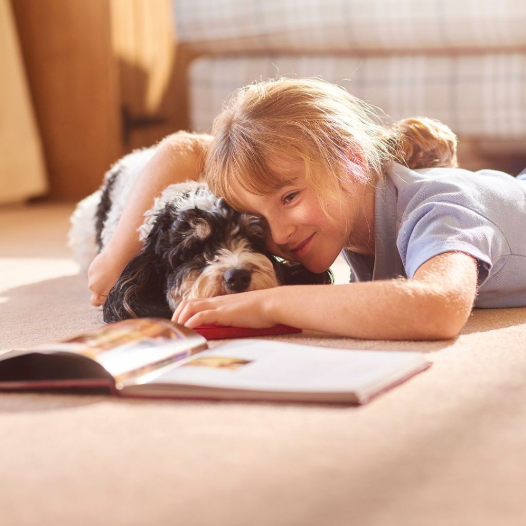A pale-skinned child with strawberry-blonde hair cuddles a shaggy black-and-white dog on the floor in front of an open picture book.