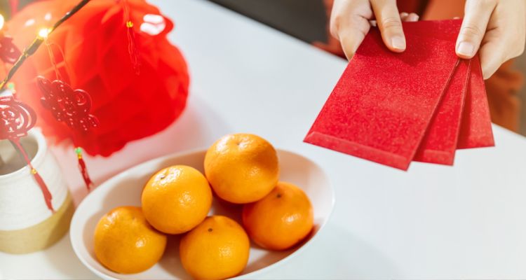 Five orange tangerines rest in a wide white dish on a white table; nearby, pale-skinned hands hold tree bright red envelopes toward the viewer; on the opposite side of the tangerines, several red, tasseled ornaments hang from a lighted artificial tree branch,
