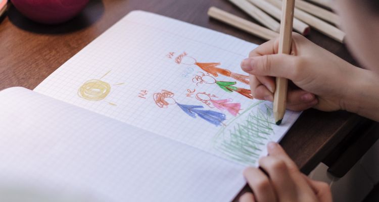 A light-skinned child uses a pencil to color in a scene in a book of graph-lined pages; already drawn are four figures with the labels "Ma," "Me," "Bro," and "Pa."