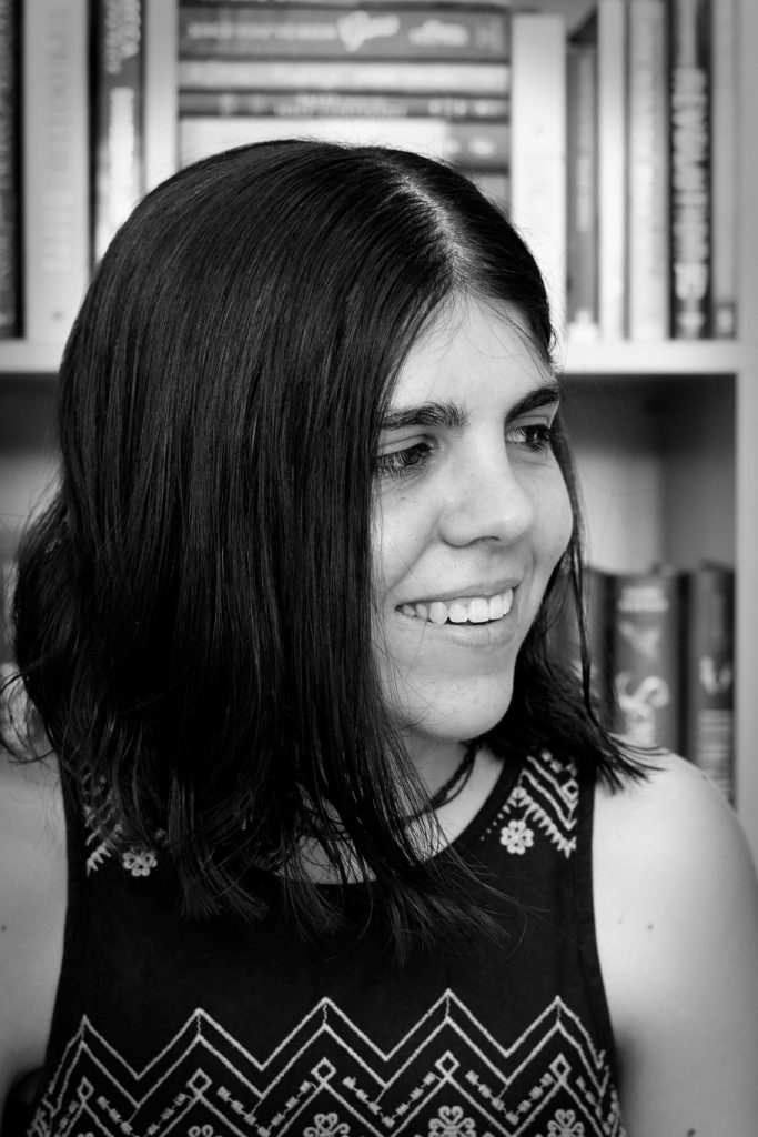 Black and white photo of CG Drews. They wear a black and white shirt and look to the right of the camera and smile.