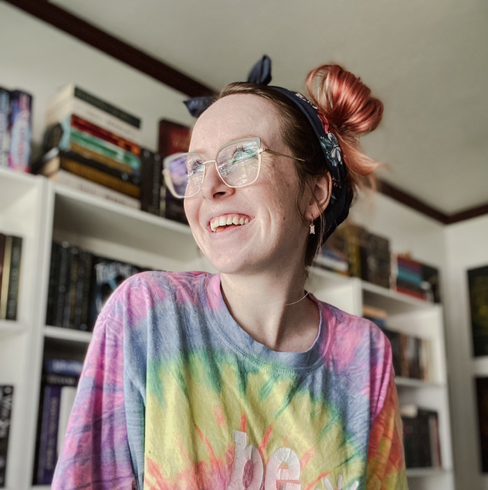 Photo of Erin K. Larson-Burnett. She wears a tie-dye shirt and is smiling to the left side of the camera with her bookshelves behind her.