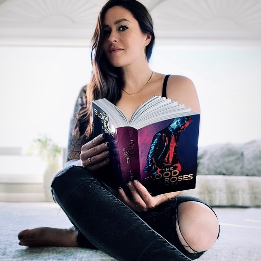 Author Callie Hart positing with her dark brown hair to the side, black jeans with a rip in the left knee, and holding her book, The Good Roses. The book has a dark theme, with a man in a leather jacket looking down and dark shadows on him.