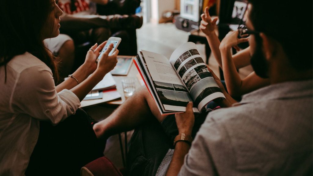 A group of individuals sit in a circle around a living room. The figure in front of the camera flips through a book. A person to the left holds their phone.
