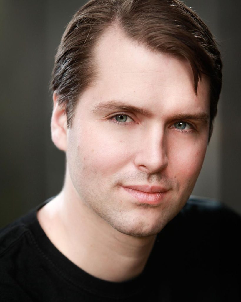 A headshot of a man with light skin, blue-green eyes, and short brown hair parted on the side. He is smiling slightly and is wearing a dark t-shirt over a gray background.