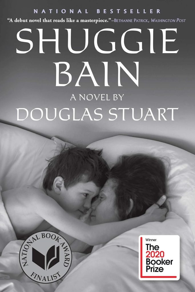 White text reads "Shuggie Bain" over a black and white photo, followed by smaller white text that reads "A Novel by Douglas Stuart". The photo background depicts a mother and a young boy, her child, touching foreheads and looking closely at each other while wrapped in a blanket and surrounded by pillows.