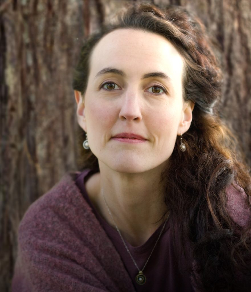 A woman with light skin, brown eyes, and dark brown wavy hair that is medium-length with highlights. She wears a purple sweater, round silver earrings and pendant, and is smiling slightly in front of a wooden background.