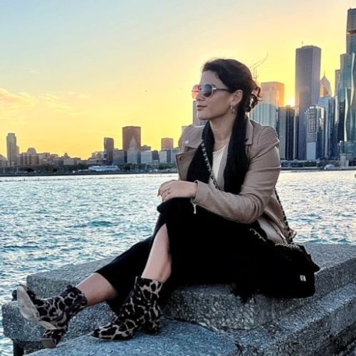 A dark haired white woman sitting on concrete wall along a waterway with a cityscape in the background at sunset.