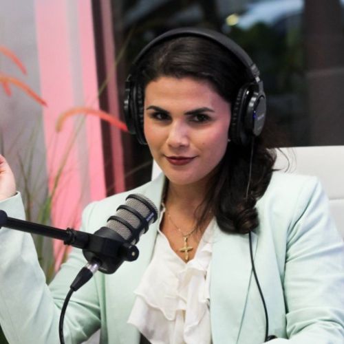 Sofia Wardell, a white woman smiling coyly at the camera sitting in a white desk chair with audio equipment set up for a podcast.