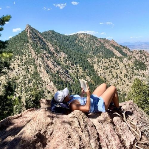 Sofia Wardell, a White woman is laying atop a boulder reading a book with a mountain in the background.