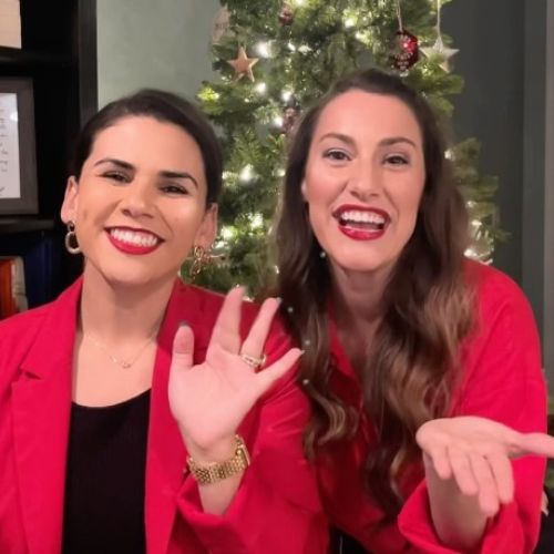 Two white women smiling and waving at the camera dressed in red blazers before a Christmas Tree.