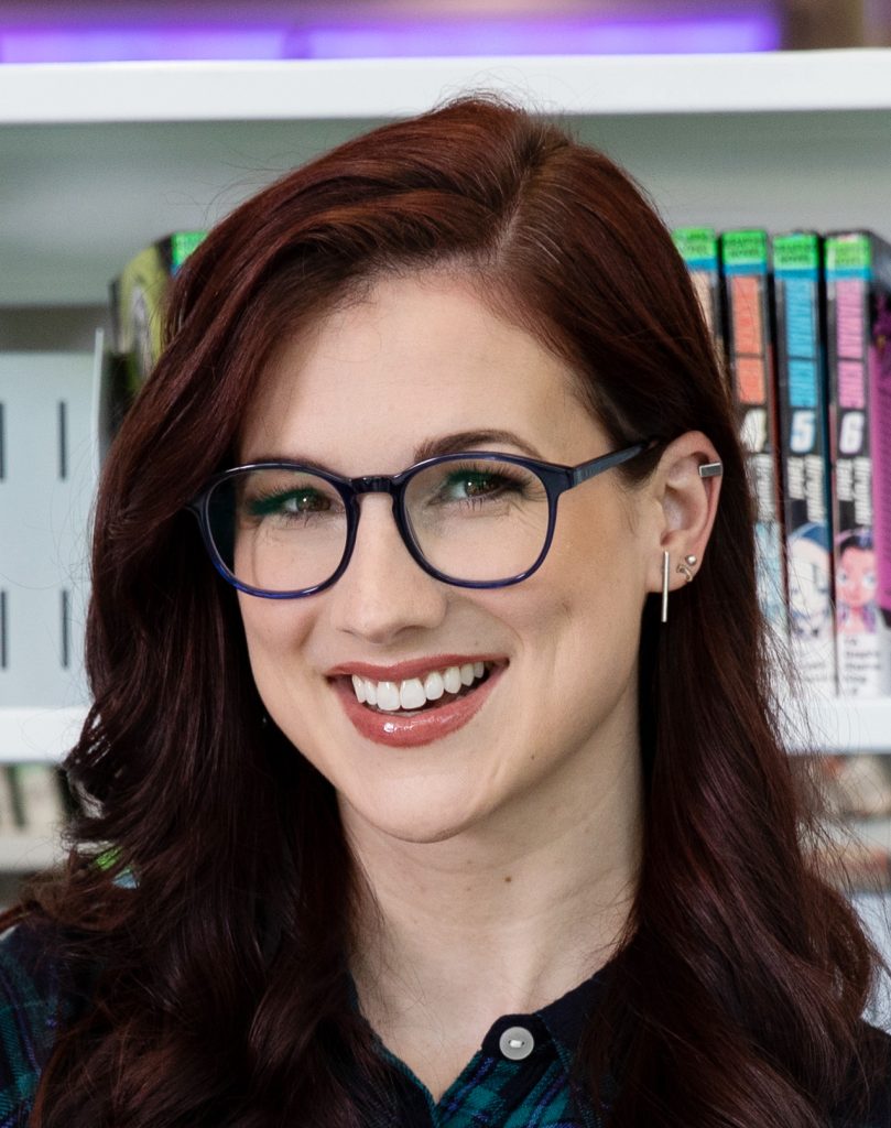 A woman looking at the camera and smiling. She has red hair and wears black frame glasses. She is standing in front of a bookshelf.