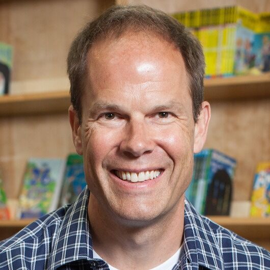 A white man with short brown hair and wearing a blue0checked shirt smiles at the viewer; the background is bookshelves containing children's books.