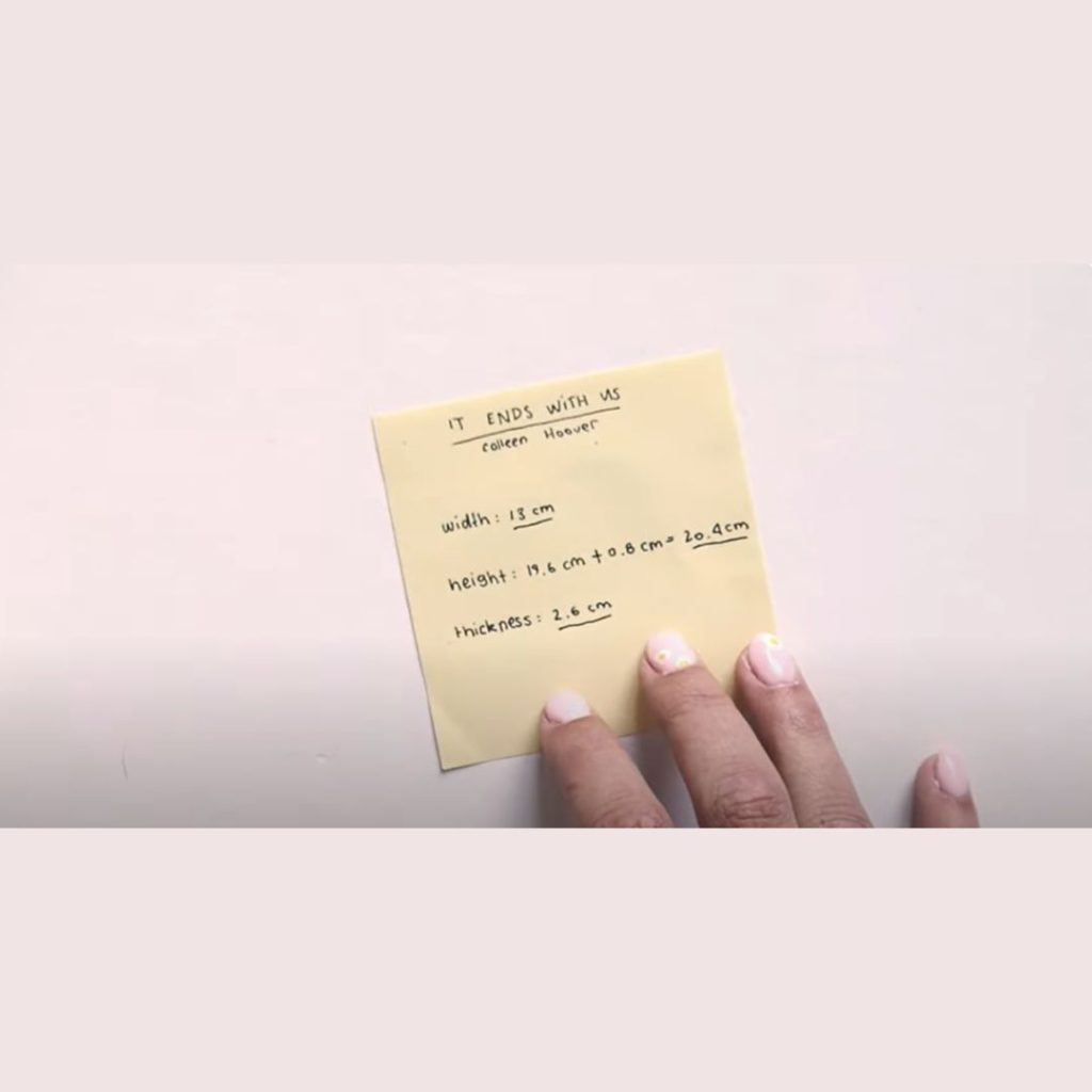 woman's hand holding down a yellow post it note that says it ends with us, colleen hoover, width is thirteen centimeters, height is nineteen point six centimeters plus zero point eight centimeters equals twenty point four centimeters and thickness is two point six centimeters