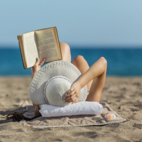 girl laying on the beach with the blue ocean in front of her while she's holding onto her hat and reading a book with one hand