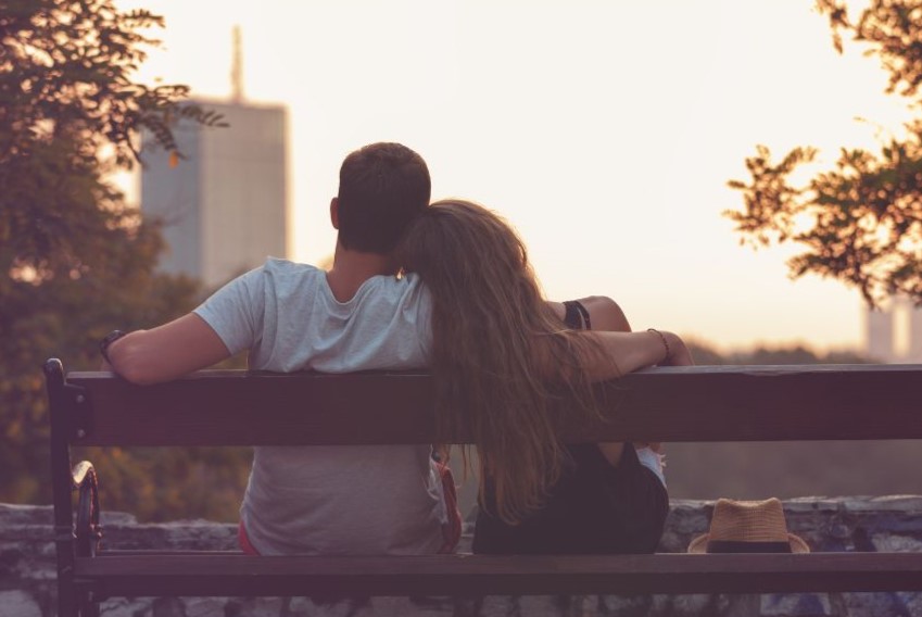 A couple sitting outside on a bench with the woman leaning her head on the man's shoulder