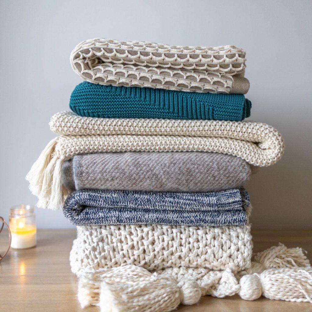 Tan, white, and blue blankets stacked on top of each other on a wood floor with a lit candle in the back