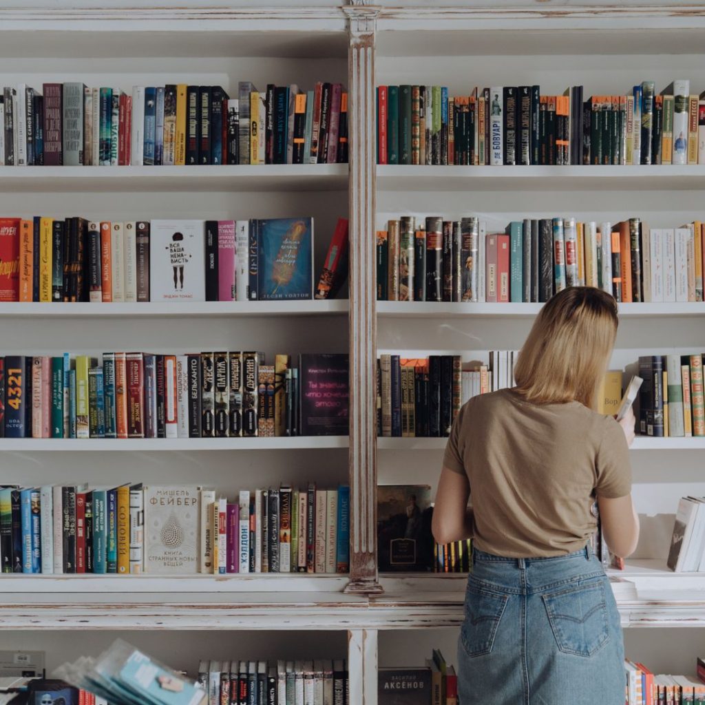 White weathered bookshelves full of different colored books with a girl in jeans and a brown shirt tilting her head to the side and picking up one of the books.