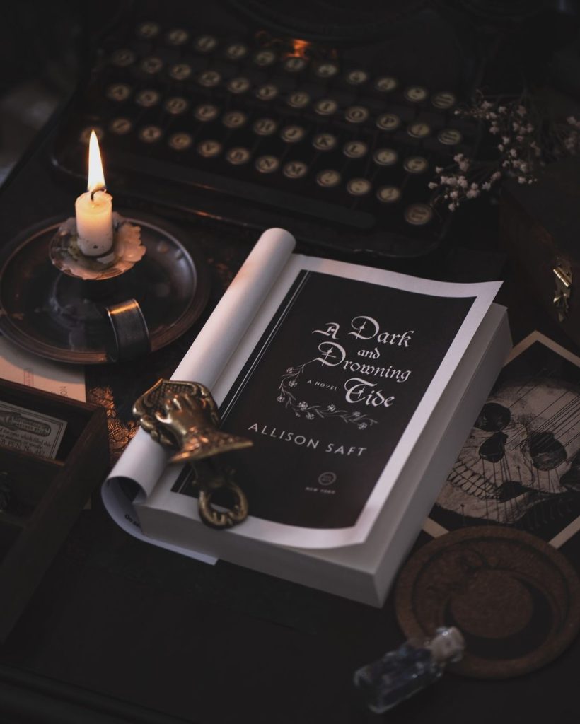 A paperback book sits open to page one with a gold trinket holding it in place. A lit candle is to the left of the book. There is a typewriter in the background.