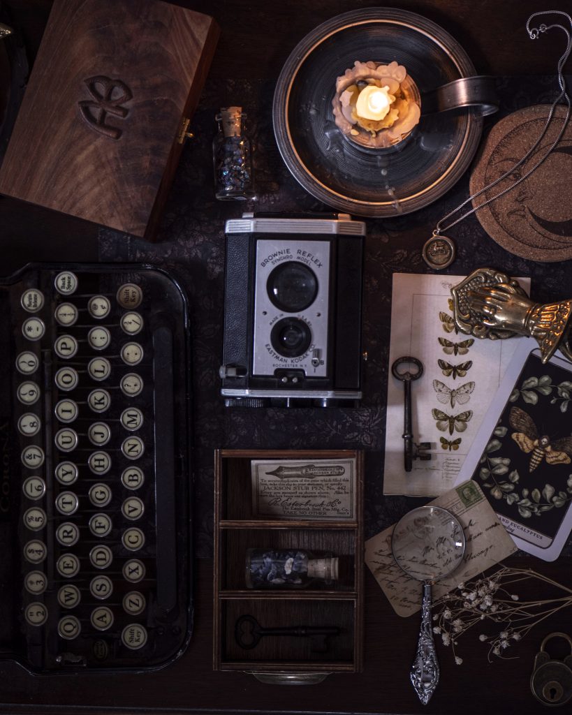 A collection of different things, which includes a camera, a key, butterflies, a pendant, a lit candle, dried flowers, and a typewriter