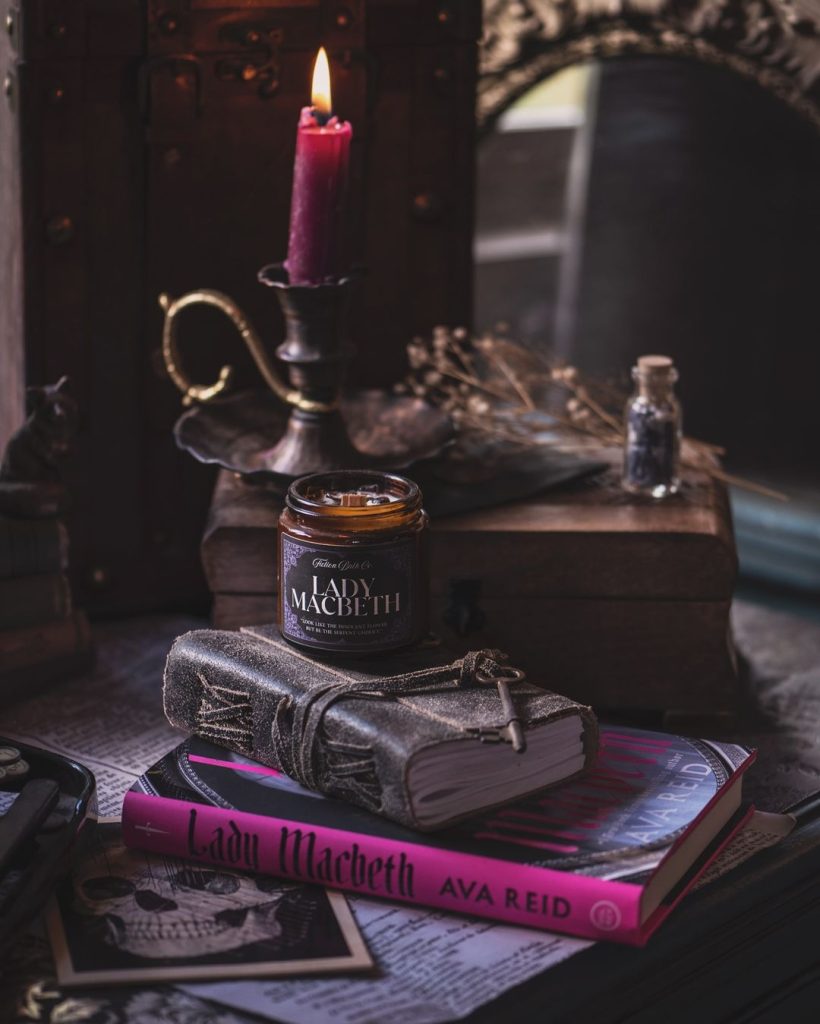 A book with a pink spine, a leather bound book, and a candle. In the background, there is a lit candle and. a glass vial.