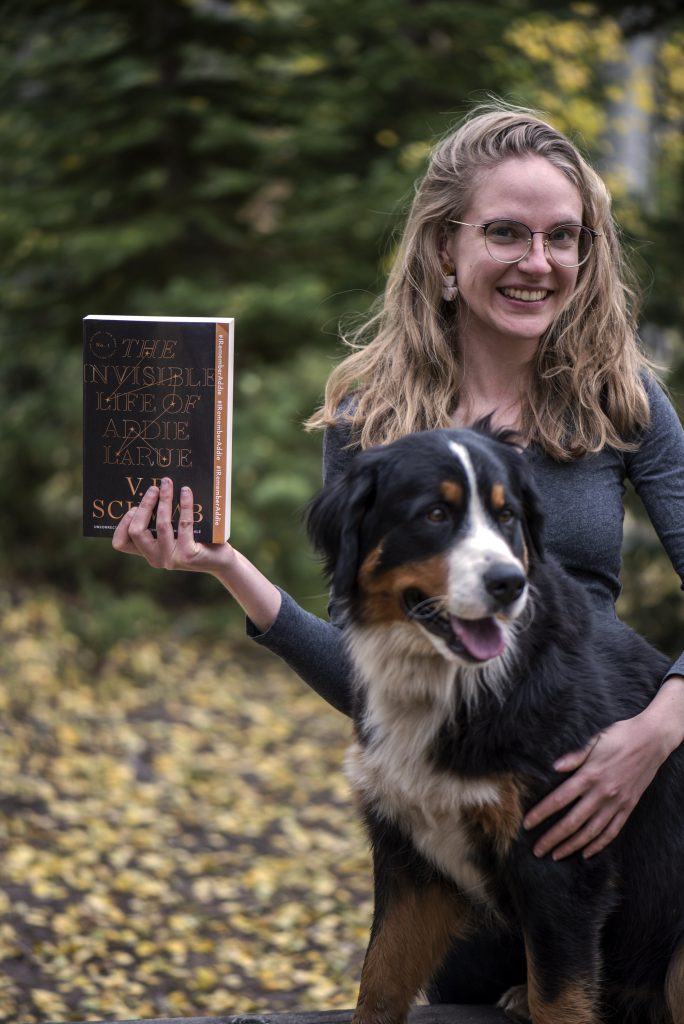 Amanda stands outside with one arm around her dog and the hand holding up a book. In the background, there are green trees and the ground is covered in yellow and brown leaves.