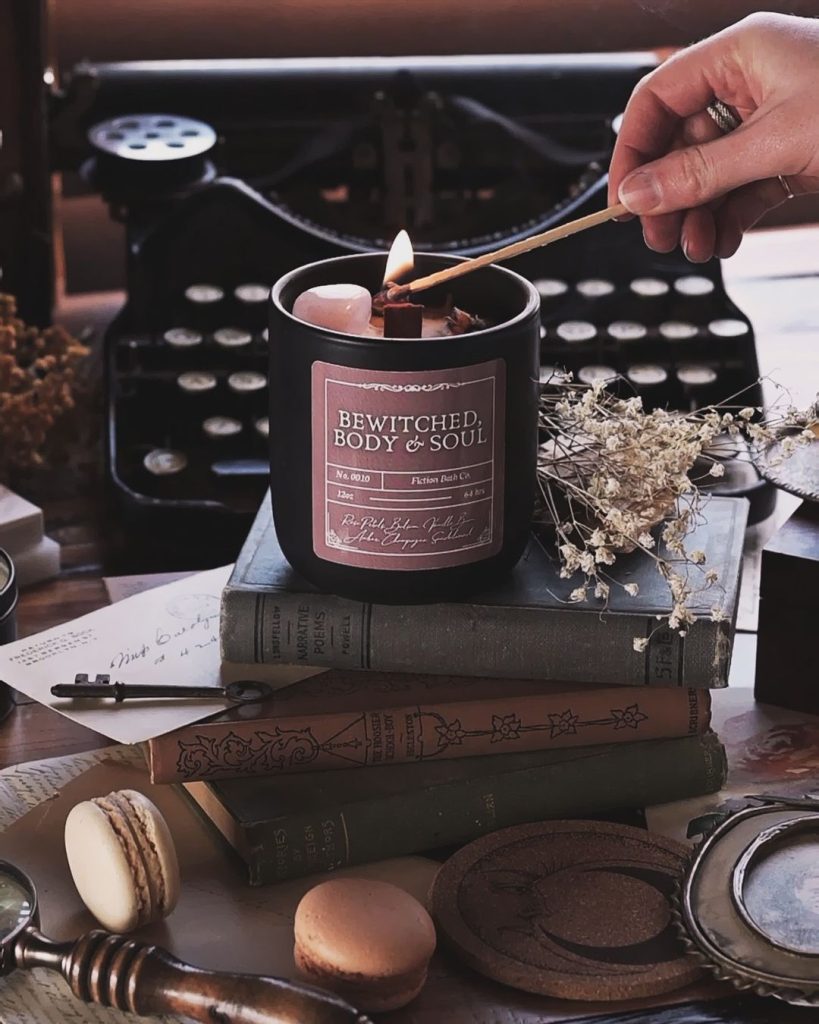 A hand lights a candle. The candle is sitting on a stack of books. Other items laying around the candle include some dried flowers, a key, macarons, an envelope, and a typewriter.