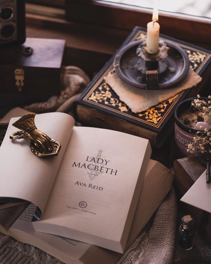 An open book sits on top of another open book. A gold trinket holds it open. A lit candle sits on top of a book in the background.