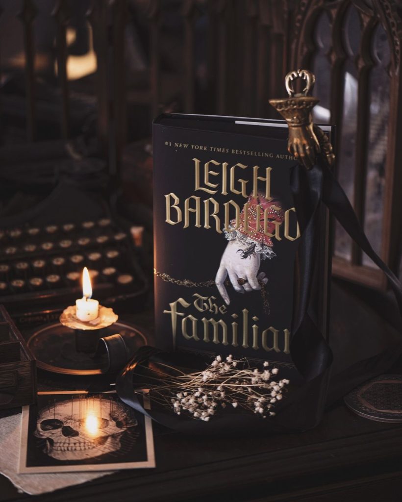 A book propped up on a table. Besides the book is a lit candle with dried flowers and a postcard with a skull. A typewriter is in the background.