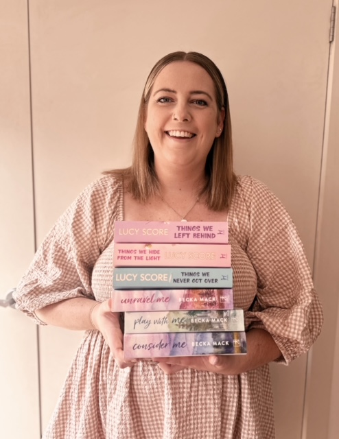 Booksagrammer Tay posed in front of a beige wall holding a stack of books with pastel covers. Tay is pictured wearing a plaid dress.