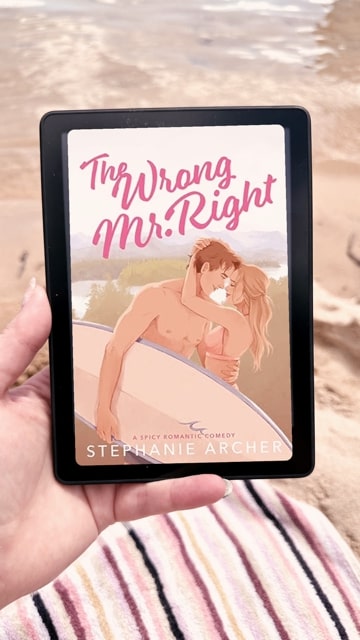 Tay holding her Kindle with sand behind it. The Kindle is displaying the cover of the book "The Wrong Mr.Right," which features a cartoon drawing of a man embracing a woman while also holding a surf board.