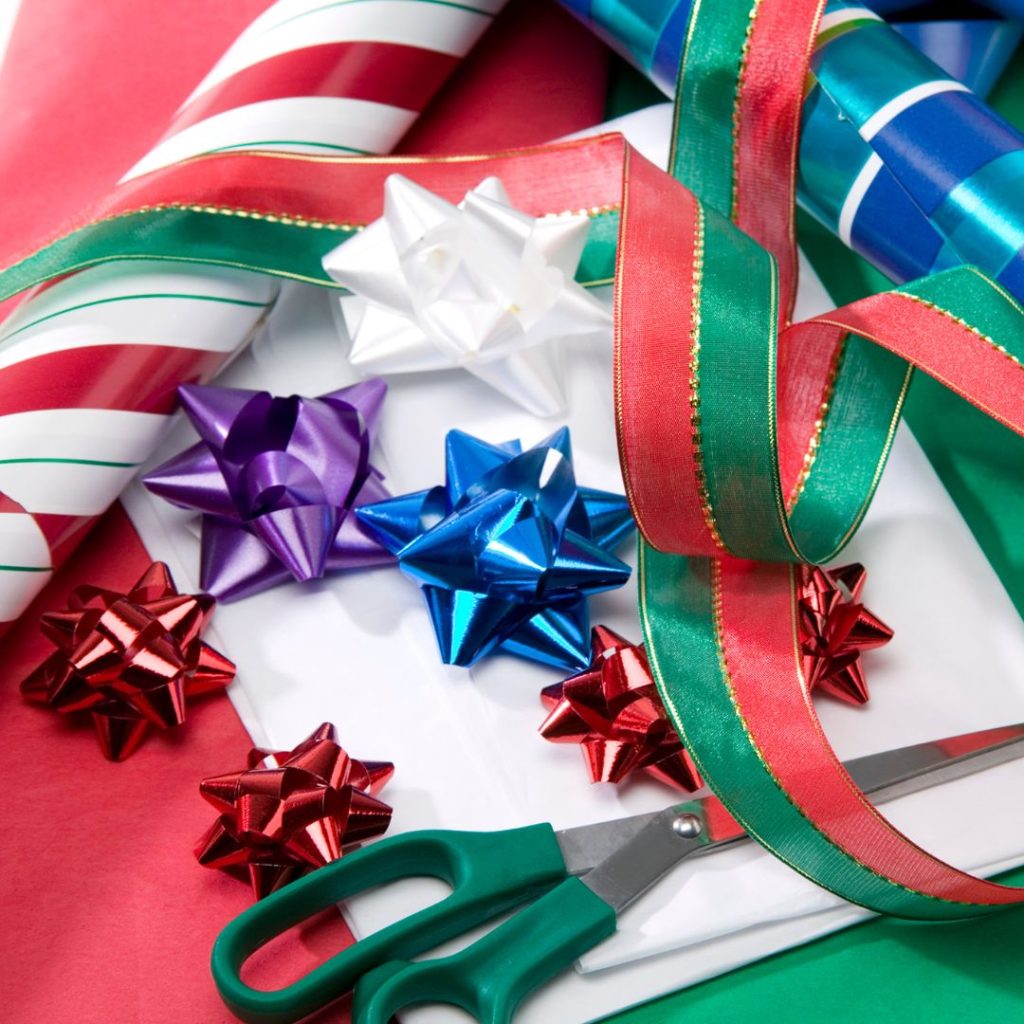 Two rolls of wrapping paper: one with red, white, and green stripes and one with blue, green, and white stripes. There are green and red ribbons and colorful bows strewn about with green-handled scissors.