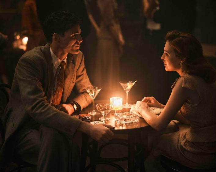A man in a vintage suit sits across a small table from a young woman in a vintage dress.