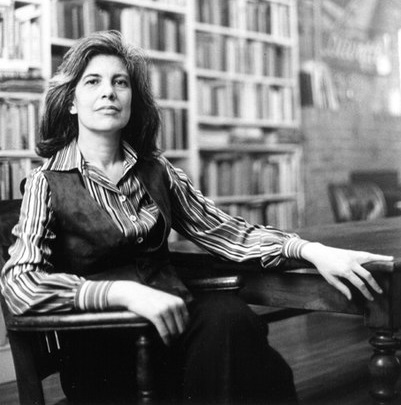 Black-and-white portrait of Susan Sontag seated at a wooden desk, surrounded by bookshelves. Photo taken by Lynn Gilbert (1979).